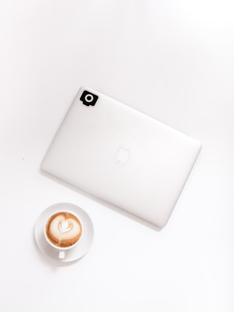 A minimalistic setup featuring a closed laptop placed on a white surface with a small black sticker on its corner. Adjacent to it is a cup of cappuccino with heart-shaped latte art.