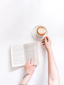 A close-up of hands holding a steaming cup of coffee beside the book on a minimalist white desk.