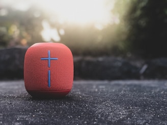 A red portable speaker with a mesh surface and a prominent plus sign on its front is placed on a textured surface. Soft, natural light creates a serene atmosphere with blurred greenery in the background.