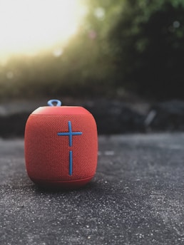 A red portable Bluetooth speaker with a prominent blue cross design resting on a textured concrete surface. The background is softly blurred, featuring natural greenery and light coming through, creating a pleasant bokeh effect.