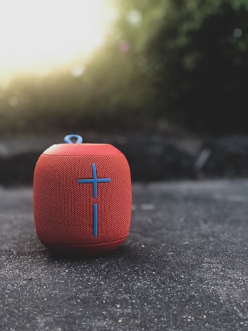 A red portable Bluetooth speaker with a prominent blue cross design resting on a textured concrete surface. The background is softly blurred, featuring natural greenery and light coming through, creating a pleasant bokeh effect.