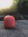 A red portable Bluetooth speaker with a prominent blue cross design resting on a textured concrete surface. The background is softly blurred, featuring natural greenery and light coming through, creating a pleasant bokeh effect.