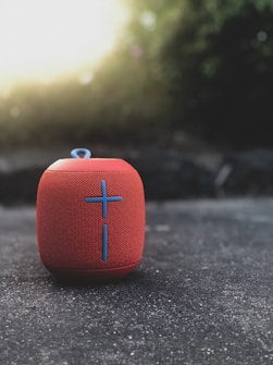 A red portable Bluetooth speaker with a prominent blue cross design resting on a textured concrete surface. The background is softly blurred, featuring natural greenery and light coming through, creating a pleasant bokeh effect.