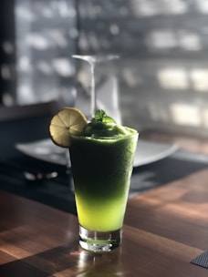 A refreshing green smoothie topped with a lemon slice and garnished with mint. The glass sits on a wooden table with soft dappled sunlight creating a cozy atmosphere. In the background, there is a blurred dining setup indicating a casual dining setting.