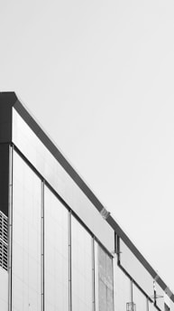 A modern building facade with a minimalist design, featuring clean lines and a monochrome color scheme. The structure has large rectangular panels and subtle architectural details.