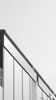 A modern building facade with a minimalist design, featuring clean lines and a monochrome color scheme. The structure has large rectangular panels and subtle architectural details.