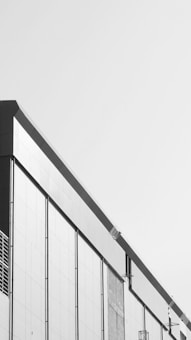 A modern building facade with a minimalist design, featuring clean lines and a monochrome color scheme. The structure has large rectangular panels and subtle architectural details.