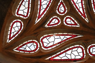 Intricate stained glass design featuring teardrop-shaped panes bordered by vibrant red outlines and interspersed with clear glass. The arrangement forms a symmetrical and harmonious pattern, set against a wooden or stone framework.