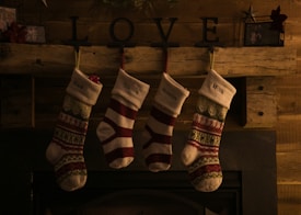 Four Christmas stockings hang from a wooden mantle in a dimly lit room. Each stocking has a name on it: Dad, Tristan, Madelyn, and Mom. Above the stockings, the word LOVE is displayed using metal letters. The mantle is decorated with small, festive items, enhancing the cozy holiday atmosphere.