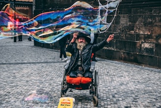 An individual in a wheelchair is creating large, colorful bubbles on a cobblestone street. The person, wearing a hat and a leather jacket, appears joyful and engaged in the activity. In the foreground, a caution sign indicates a wet floor.