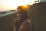 A person with closed eyes holds a peaceful expression, backlit by the setting sun. The golden sunlight creates a warm halo effect around the head, with long dark hair gently flowing. The background features blurred greenery, suggesting a natural outdoor setting, and a structure is visible in the distance on the right.