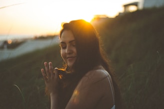 A person with closed eyes holds a peaceful expression, backlit by the setting sun. The golden sunlight creates a warm halo effect around the head, with long dark hair gently flowing. The background features blurred greenery, suggesting a natural outdoor setting, and a structure is visible in the distance on the right.