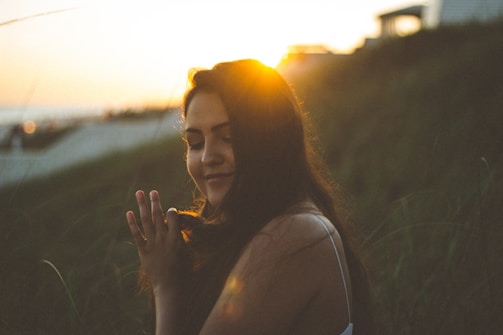 A person with closed eyes holds a peaceful expression, backlit by the setting sun. The golden sunlight creates a warm halo effect around the head, with long dark hair gently flowing. The background features blurred greenery, suggesting a natural outdoor setting, and a structure is visible in the distance on the right.