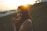 A person with closed eyes holds a peaceful expression, backlit by the setting sun. The golden sunlight creates a warm halo effect around the head, with long dark hair gently flowing. The background features blurred greenery, suggesting a natural outdoor setting, and a structure is visible in the distance on the right.