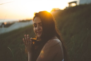 A person with closed eyes holds a peaceful expression, backlit by the setting sun. The golden sunlight creates a warm halo effect around the head, with long dark hair gently flowing. The background features blurred greenery, suggesting a natural outdoor setting, and a structure is visible in the distance on the right.