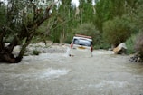 A safari jeep crossing a shallow river with hippos and elephants nearby.