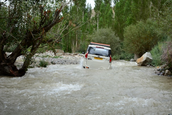 A 4x4 vehicle crossing a shallow river with rocky banks under a bright blue sky.