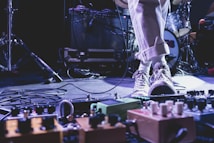 A musician's foot is stepping on various guitar effects pedals on a stage. The musician is wearing rolled-up beige pants and canvas sneakers. Cables are scattered on the floor, and music equipment like an amplifier and drum set are visible in the background.