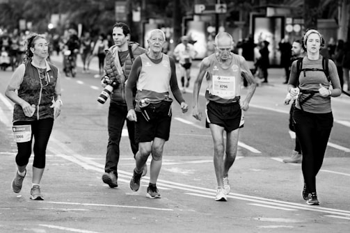 A group of people, including runners and a photographer, move along a city street during a public running event. Some participants wear race bibs, while a young woman appears to jog beside them. One man carries a professional camera, suggesting a documentative purpose. The background features blurred spectators and elements of urban scenery.