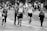 A group of people, including runners and a photographer, move along a city street during a public running event. Some participants wear race bibs, while a young woman appears to jog beside them. One man carries a professional camera, suggesting a documentative purpose. The background features blurred spectators and elements of urban scenery.