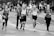 A group of people, including runners and a photographer, move along a city street during a public running event. Some participants wear race bibs, while a young woman appears to jog beside them. One man carries a professional camera, suggesting a documentative purpose. The background features blurred spectators and elements of urban scenery.