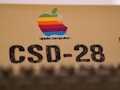 A vintage Apple Computer logo with a multicolored apple shape is placed on a beige surface. Below the logo, the text 'CSD-28' is printed in large, bold black letters.