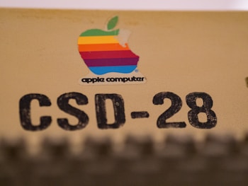 A vintage Apple Computer logo with a multicolored apple shape is placed on a beige surface. Below the logo, the text 'CSD-28' is printed in large, bold black letters.