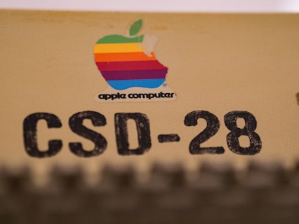A vintage Apple Computer logo with a multicolored apple shape is placed on a beige surface. Below the logo, the text 'CSD-28' is printed in large, bold black letters.