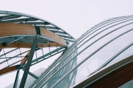 A modern architectural structure featuring a combination of steel and wooden beams with large glass panels, forming an intricate and curvilinear design against a pale sky.