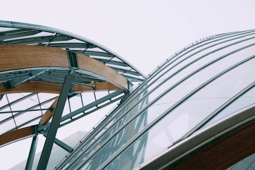 A modern architectural structure featuring a combination of steel and wooden beams with large glass panels, forming an intricate and curvilinear design against a pale sky.