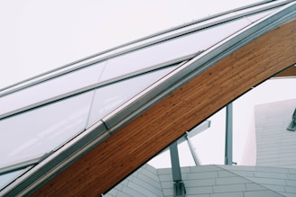 A modern architectural structure featuring a large, curved wooden beam supporting a glass roof. The structure is complemented by metal beams and a paneled facade.