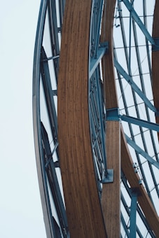Curved wooden beams are interwoven with a metallic steel framework, showcasing an architectural structure with clean geometric lines. The composition contrasts the warmth of the wood with the coolness of the steel, highlighted against a pale sky.