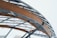 A close-up view of a modern architectural structure featuring curved wooden beams and metal supports forming an intricate geometric pattern. The background is a light, overcast sky, providing a minimalist and clean backdrop.