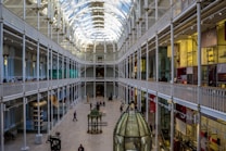 An expansive and brightly lit interior of a museum featuring high arched ceilings and multiple levels of galleries. Visitors can be seen walking along the upper balconies and exploring exhibits on the ground floor. The architecture includes ornamental designs and large glass windows allowing natural light to flood the space.