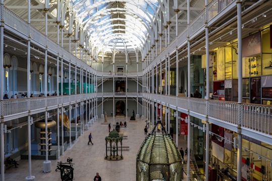 An expansive and brightly lit interior of a museum featuring high arched ceilings and multiple levels of galleries. Visitors can be seen walking along the upper balconies and exploring exhibits on the ground floor. The architecture includes ornamental designs and large glass windows allowing natural light to flood the space.