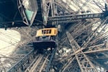 A yellow elevator car is ascending within the intricate iron lattice structure of the Eiffel Tower. The perspective from below emphasizes the complex network of beams and rivets, showcasing industrial architecture.
