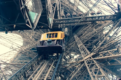 A yellow elevator car is ascending within the intricate iron lattice structure of the Eiffel Tower. The perspective from below emphasizes the complex network of beams and rivets, showcasing industrial architecture.