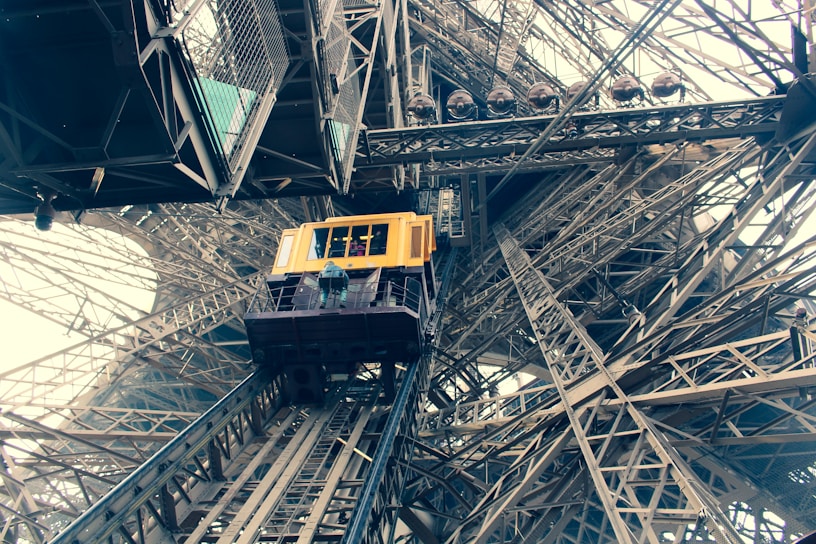 A yellow elevator car is ascending within the intricate iron lattice structure of the Eiffel Tower. The perspective from below emphasizes the complex network of beams and rivets, showcasing industrial architecture.