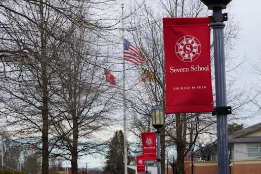 Red banners featuring the Severn School logo and the motto 'Courage to Lead' are displayed on lampposts along a pathway. Bare trees and an American flag are in the background along with several school buildings.
