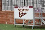 A sign with 'Severn Admirals' and 'Severn Athletics' is mounted on a wheeled, metal stand. It rests on a green, grassy surface with a brick wall in the background, and behind the wall are metal bleachers.