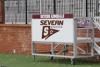 A sign with 'Severn Admirals' and 'Severn Athletics' is mounted on a wheeled, metal stand. It rests on a green, grassy surface with a brick wall in the background, and behind the wall are metal bleachers.