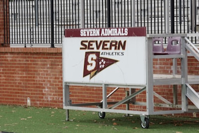 A sign with 'Severn Admirals' and 'Severn Athletics' is mounted on a wheeled, metal stand. It rests on a green, grassy surface with a brick wall in the background, and behind the wall are metal bleachers.