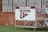 A sign with 'Severn Admirals' and 'Severn Athletics' is mounted on a wheeled, metal stand. It rests on a green, grassy surface with a brick wall in the background, and behind the wall are metal bleachers.