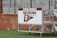 A sign with 'Severn Admirals' and 'Severn Athletics' is mounted on a wheeled, metal stand. It rests on a green, grassy surface with a brick wall in the background, and behind the wall are metal bleachers.