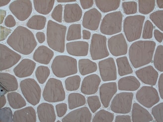 A textured stone wall pattern consisting of variously sized and shaped brown stones arranged in a mosaic with white grout lines between them.