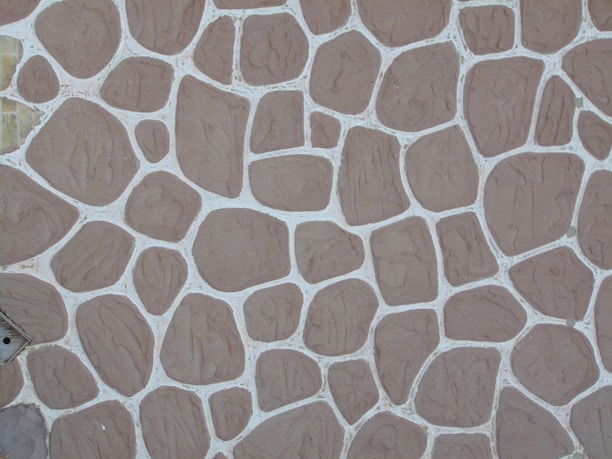 A textured stone wall pattern consisting of variously sized and shaped brown stones arranged in a mosaic with white grout lines between them.