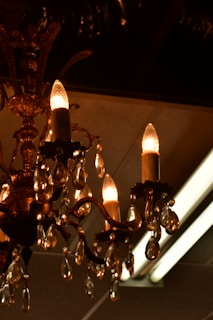 An ornate chandelier with multiple candle-like light bulbs is adorned with hanging crystal pendants. The chandelier exhibits intricate metalwork with decorative elements and a warm, amber glow from the bulbs and crystals.
