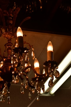 An ornate chandelier with multiple candle-like light bulbs is adorned with hanging crystal pendants. The chandelier exhibits intricate metalwork with decorative elements and a warm, amber glow from the bulbs and crystals.