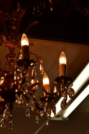 An ornate chandelier with multiple candle-like light bulbs is adorned with hanging crystal pendants. The chandelier exhibits intricate metalwork with decorative elements and a warm, amber glow from the bulbs and crystals.