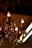 An ornate chandelier with multiple candle-like light bulbs is adorned with hanging crystal pendants. The chandelier exhibits intricate metalwork with decorative elements and a warm, amber glow from the bulbs and crystals.